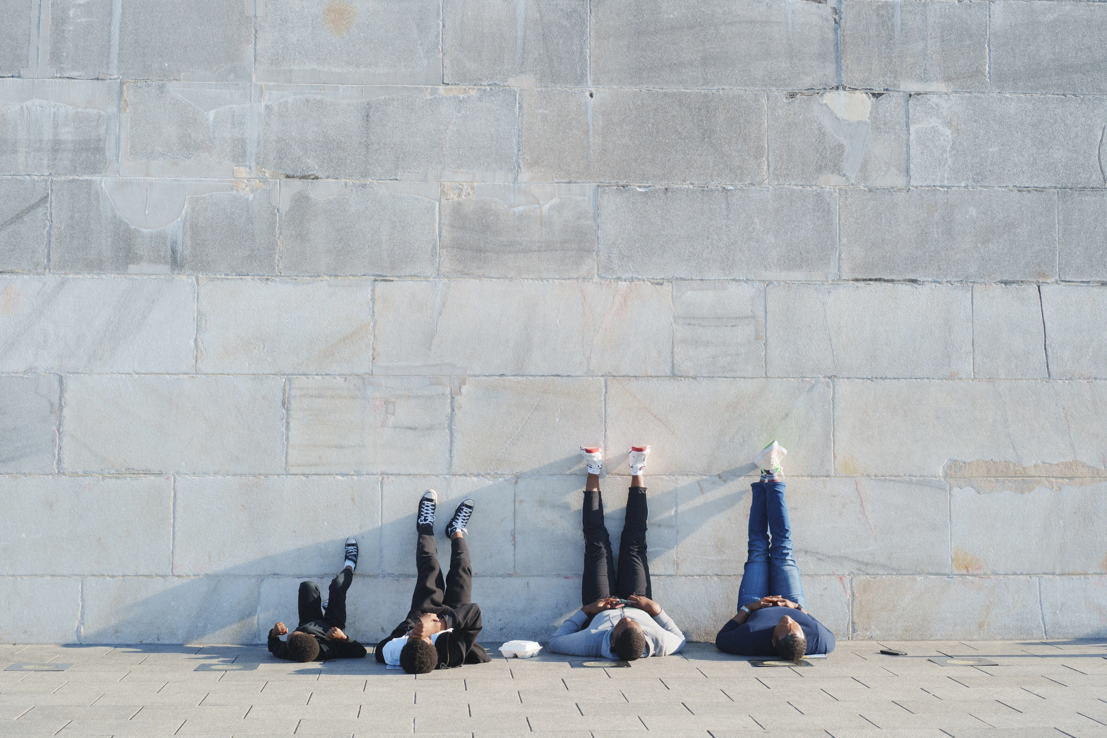 Four young people lie on their backs on a stone plaza at the foot of a massive pale monument wall, Washington, D.C.