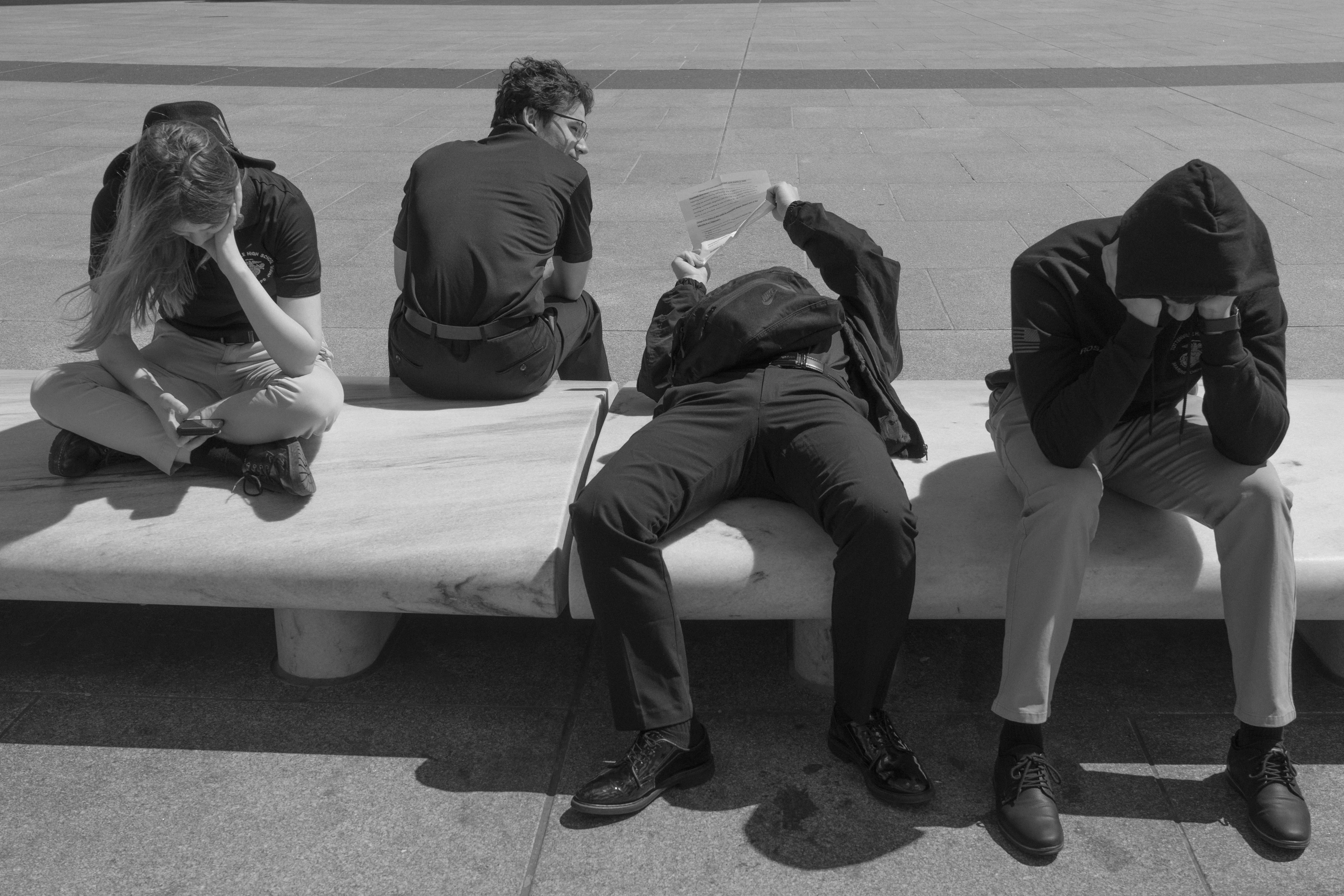 Four people seated along a long, low marble bench on a bright stone plaza in hard midday sun, Washington, D.C.
